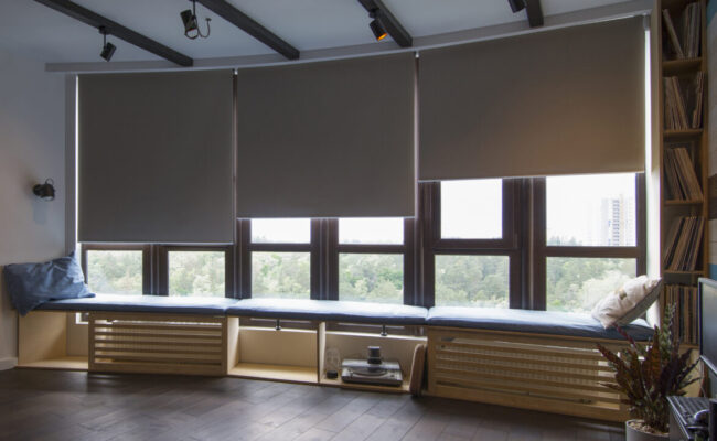 the interior. Automatic roller blinds beige color on big glass windows. Remote Control Shades are above the windosill with pillows. Summer. Green trees outside.