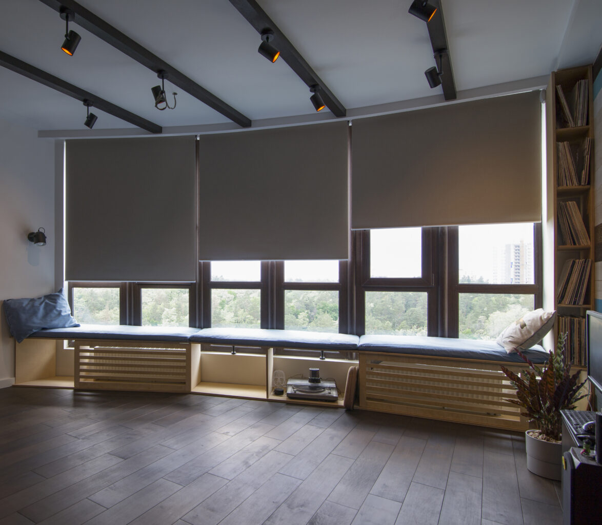 the interior. Automatic roller blinds beige color on big glass windows. Remote Control Shades are above the windosill with pillows. Summer. Green trees outside.